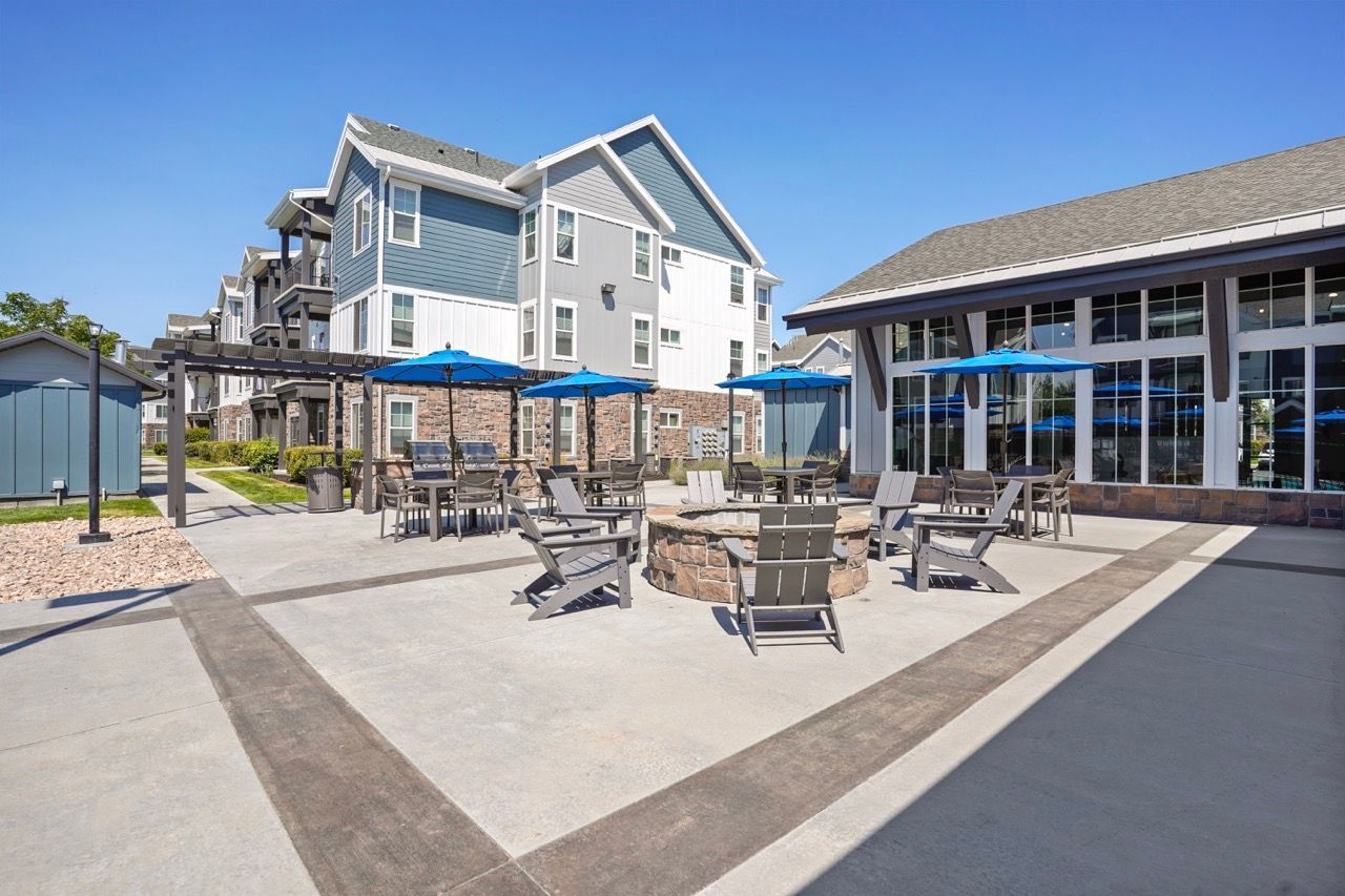 Outdoor community courtyard with seating, blue umbrellas, and a central stone fire pit near apartment buildings.