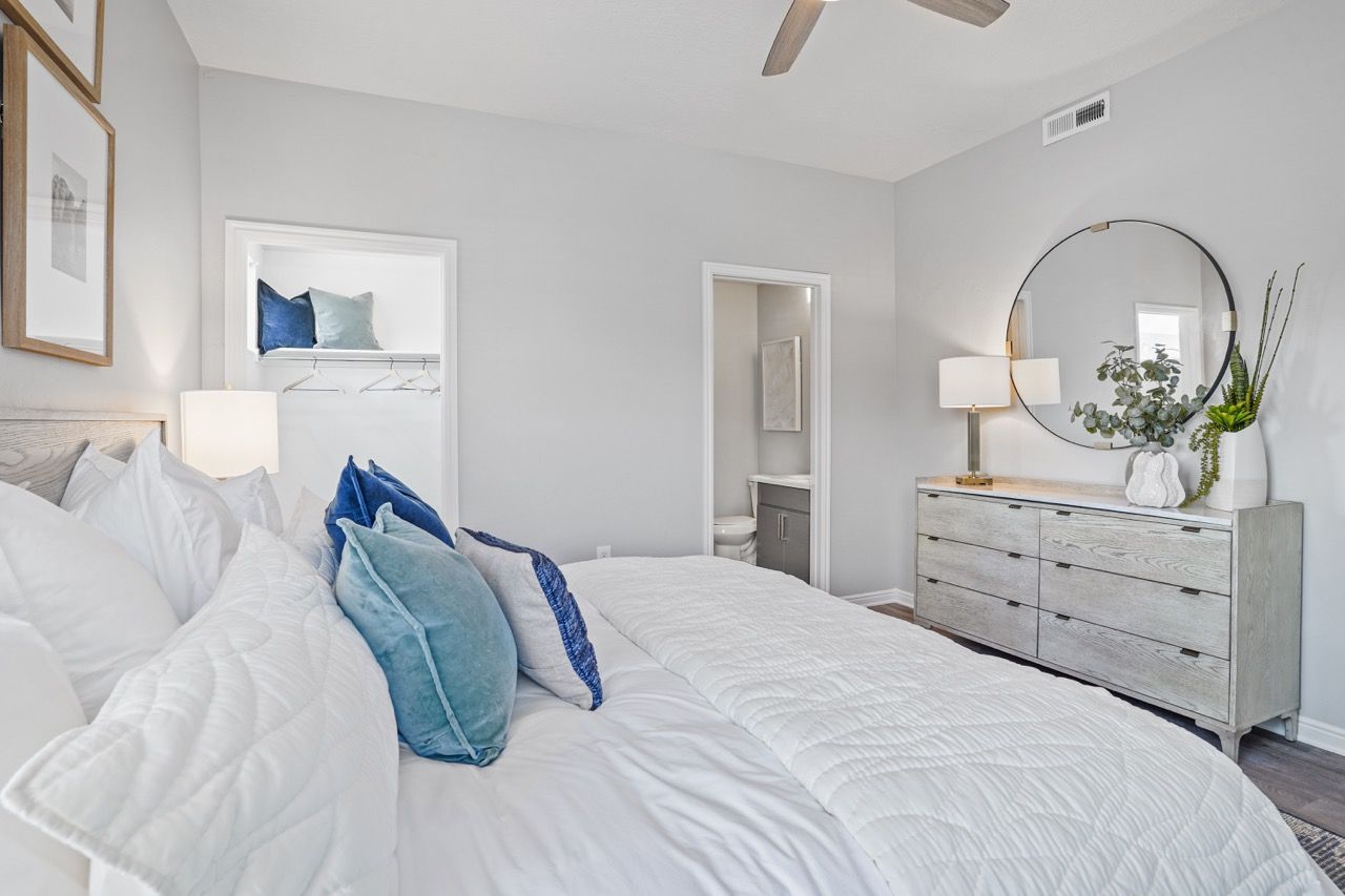 Bedroom with a large bed, white quilt, gray walls, dresser with round mirror, and plants.
