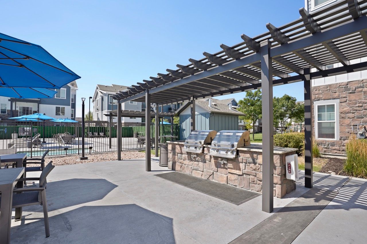 Outdoor community grilling area with stone counters under a pergola, pool and seating in background.