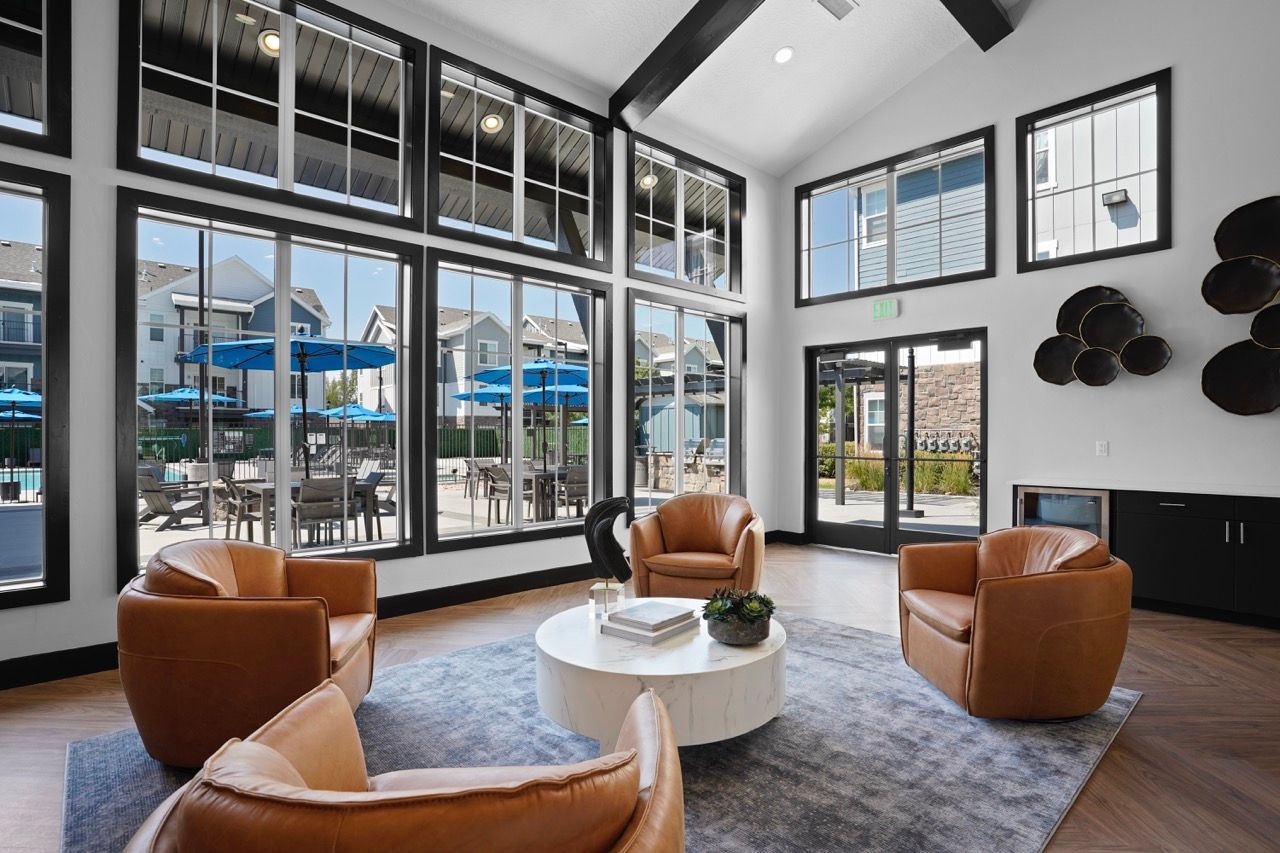 Modern resident lounge with tall windows, brown leather chairs, and a round coffee table; outdoor patio visible.