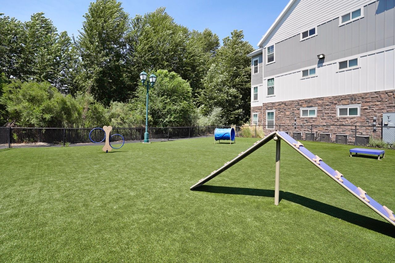 Outdoor apartment community playground with a climbing ramp, tunnel, and grassy area beside a building.