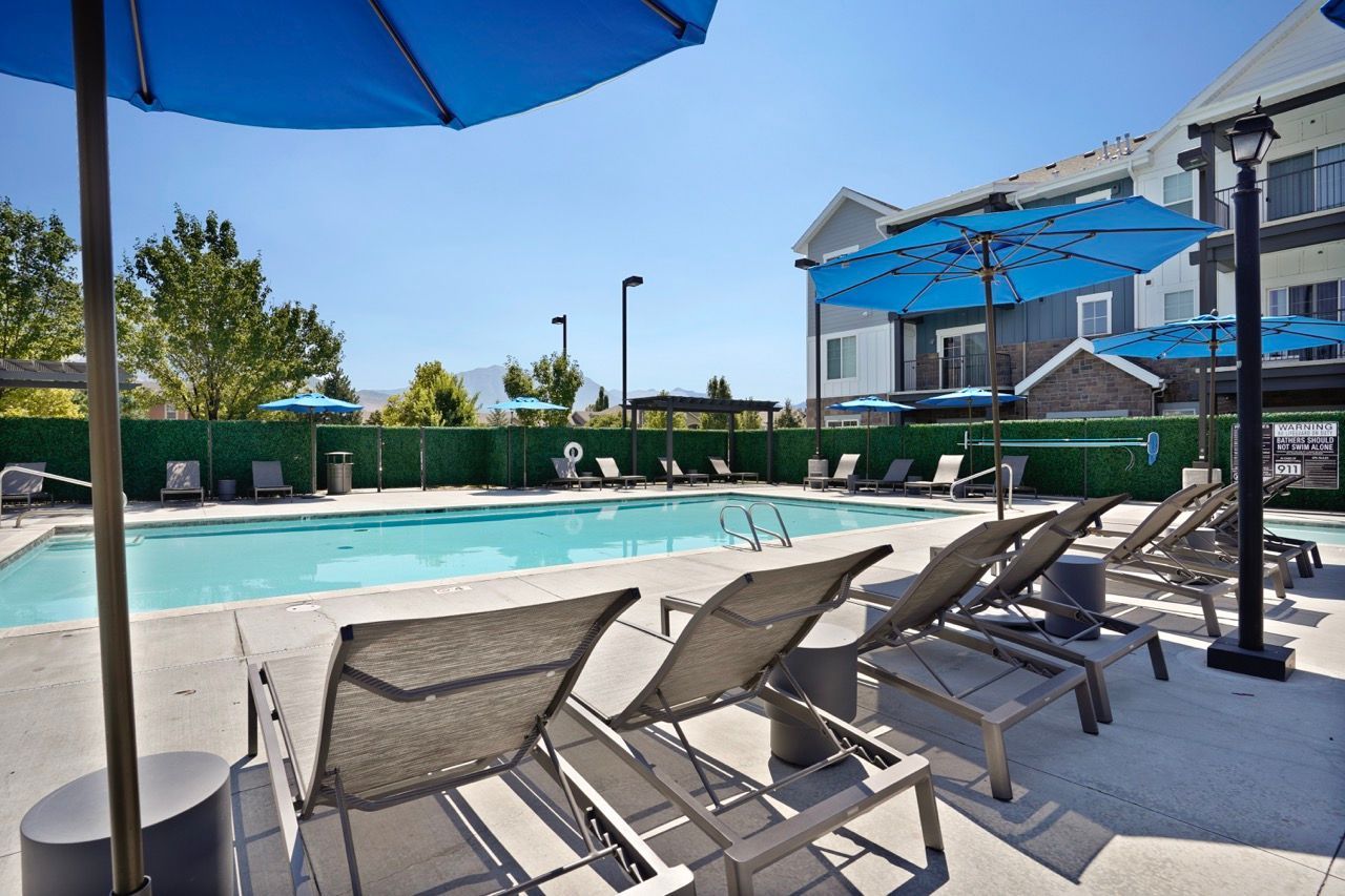 Outdoor pool area with lounge chairs, blue umbrellas, and a residential building in the background.