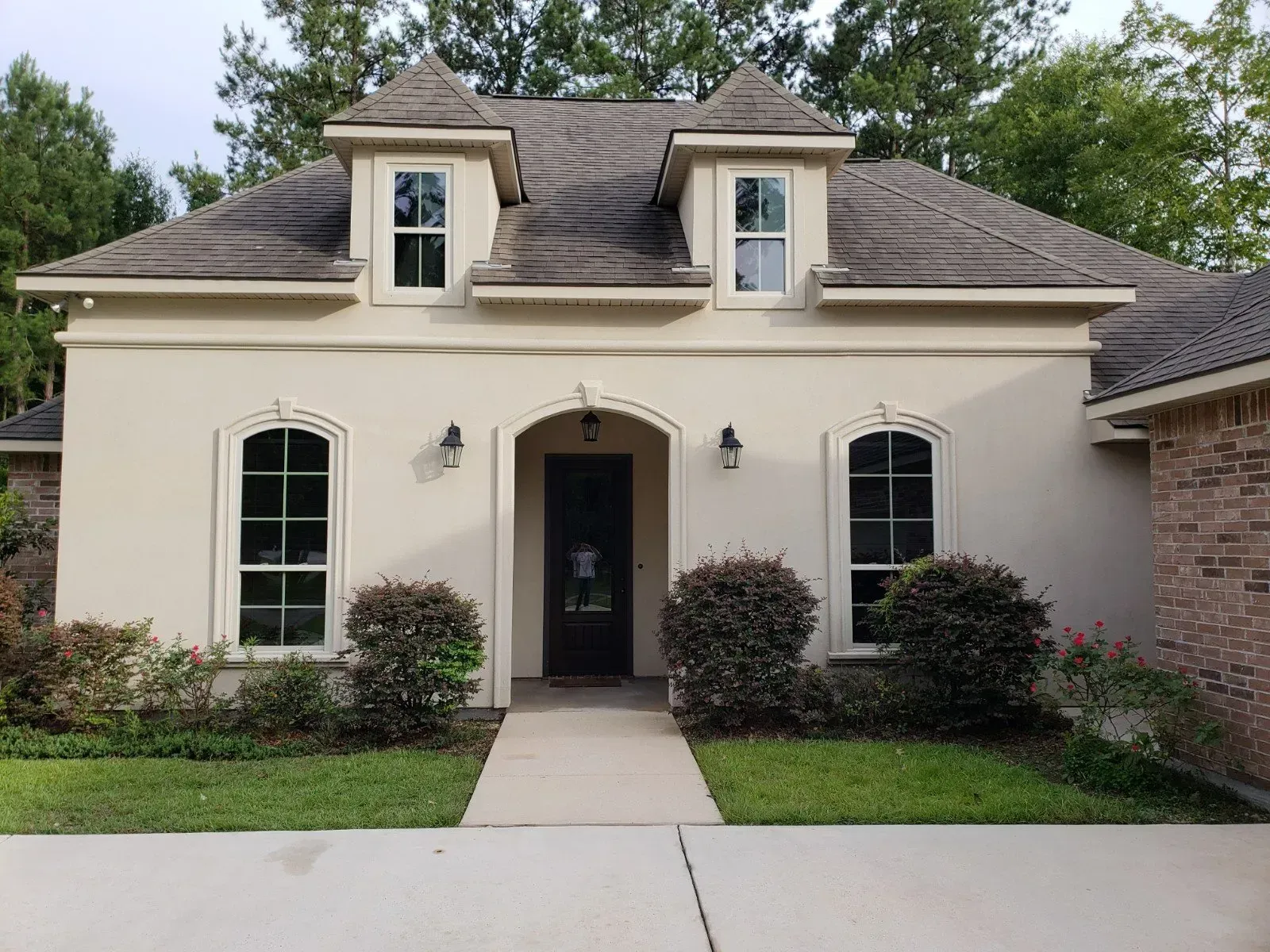 A one-story light beige stucco house with a dark front door, arched windows, and two roof dormers, set behind a lawn.