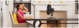 Woman sweating, fanning herself with a table fan at a table in a sunny dining room.
