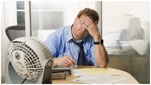 Man in office, stressed, hand on forehead, fan blowing, paperwork on desk.