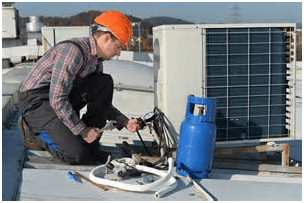 HVAC technician in orange hard hat repairs air conditioner unit on a rooftop; gas tank nearby.