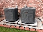 Two gray air conditioning units on concrete pads near a brick wall.