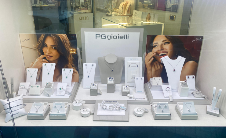 Esposizione di gioielli con collane, orecchini e anelli su supporti bianchi, corredati da foto promozionali.