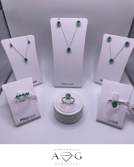 Esposizione di gioielli: collane, orecchini e anelli di smeraldi su supporti bianchi, logo AG Gioielli.