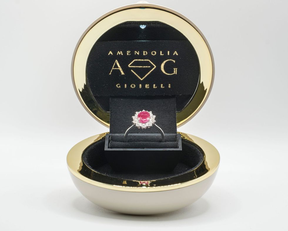 Anello in un portagioie rotondo e aperto. L'anello ha una pietra centrale rossa e pietre bianche circostanti.