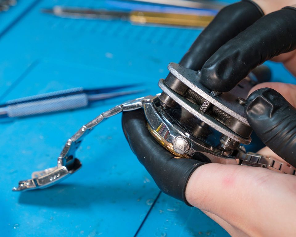 Mani in guanti neri che tengono un orologio e utilizzano uno strumento su un tappetino blu.
