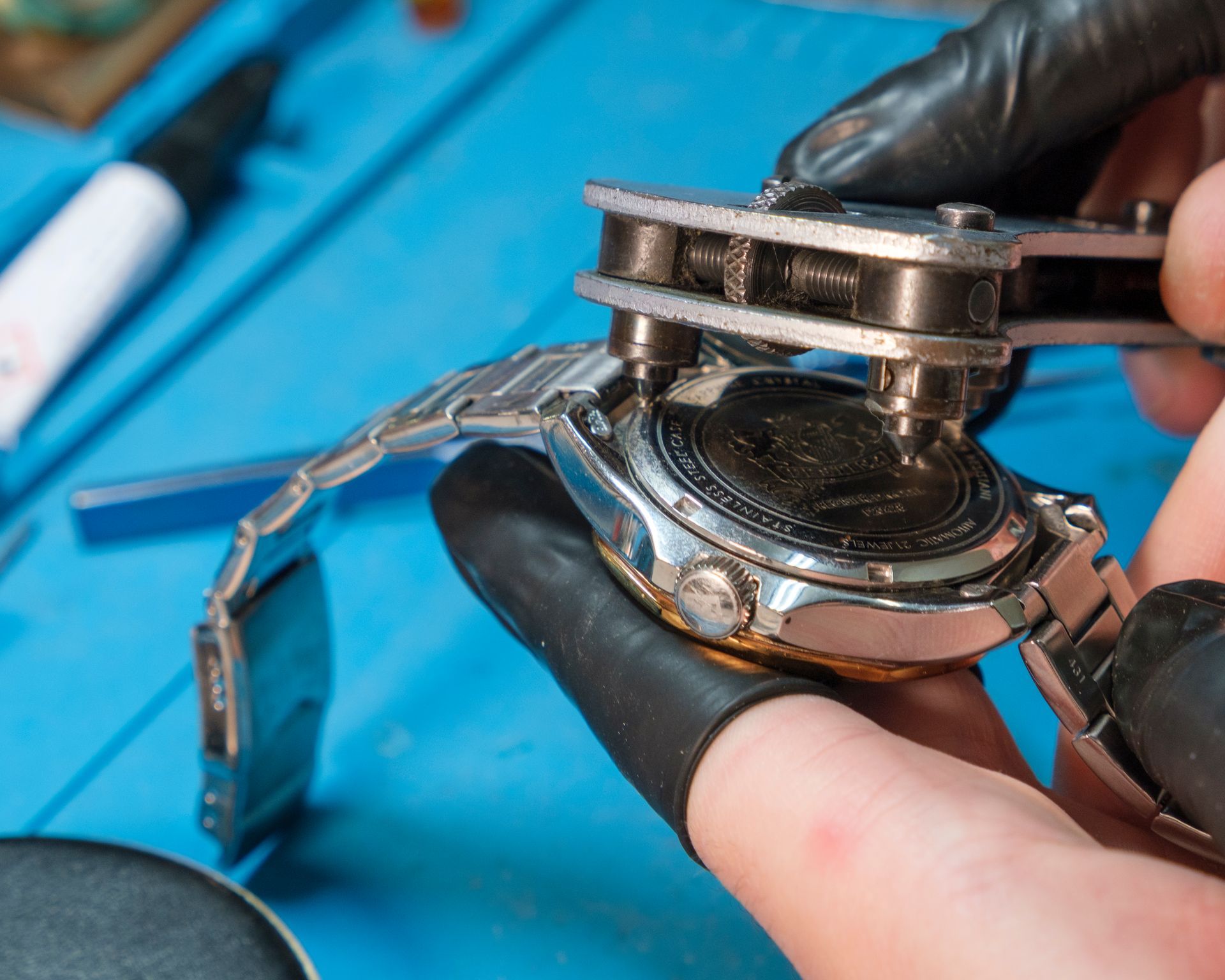 Persona che utilizza una pressa per orologi per aprire un orologio da polso. Lancette con guanti neri, tappetino blu, orologio argentato.