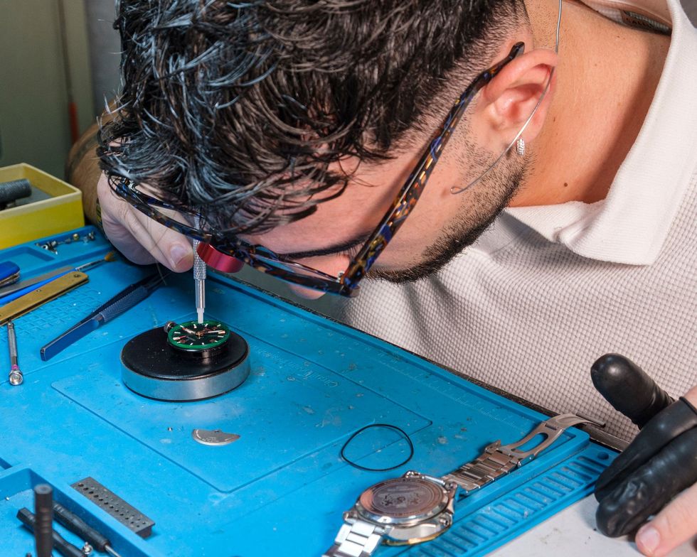 Un orologiaio lavora su un orologio, utilizzando degli strumenti su un tappetino blu.
