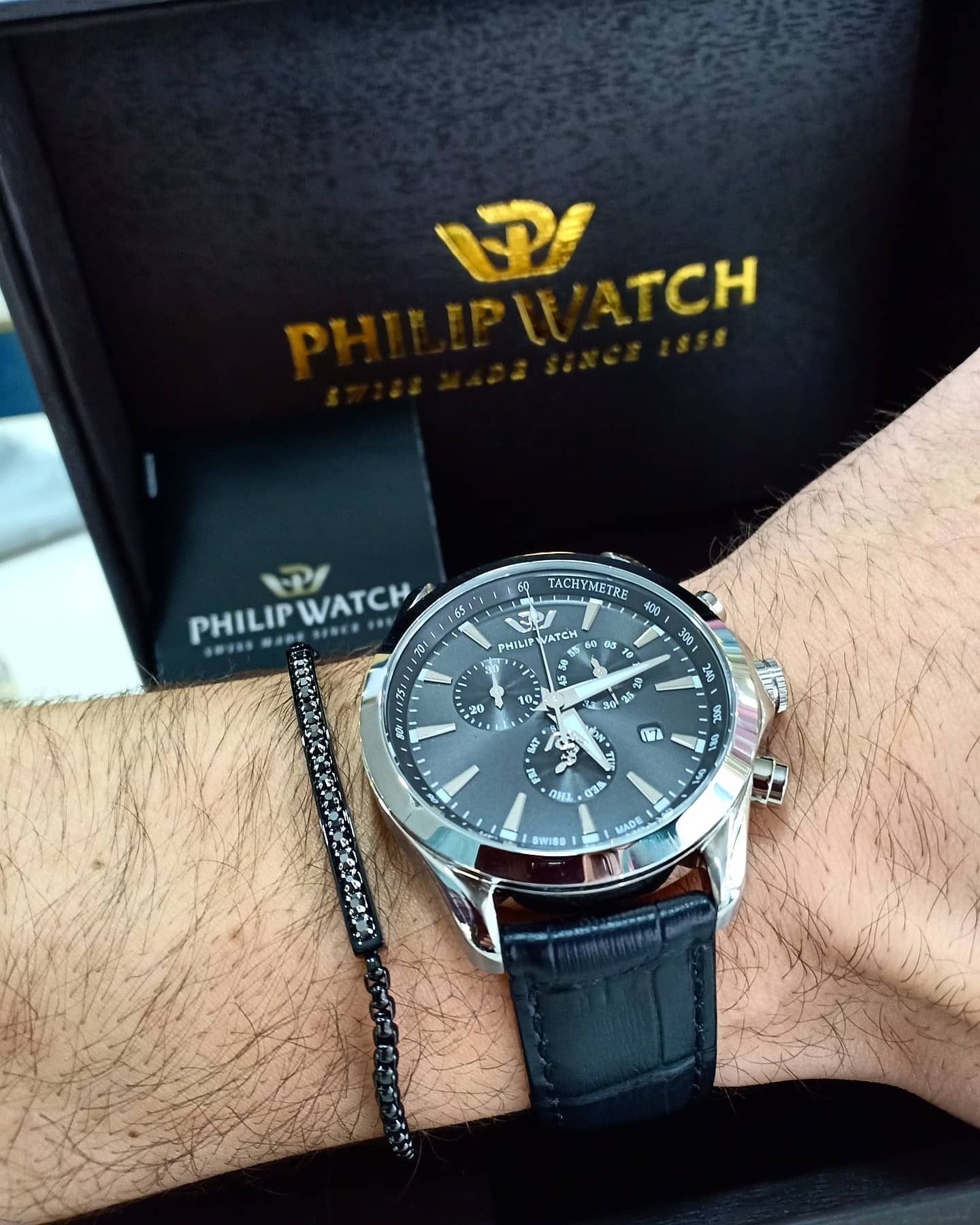 Un Philip Watch argentato al polso con cinturino in pelle nera, quadrante nero e scatola.