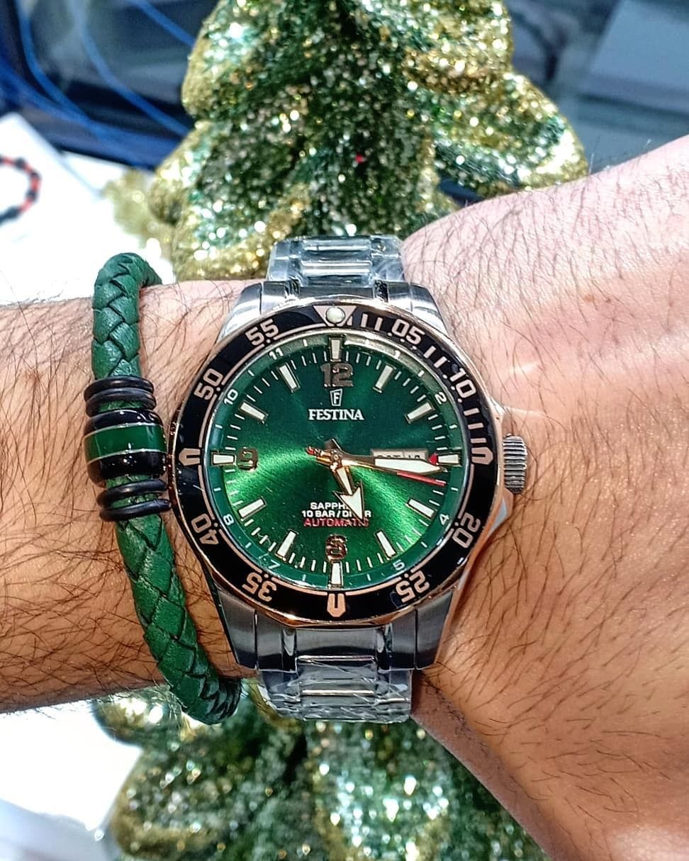 Al polso un orologio con quadrante verde e un braccialetto intrecciato verde. Entrambi gli accessori sono appoggiati a un albero di Natale.