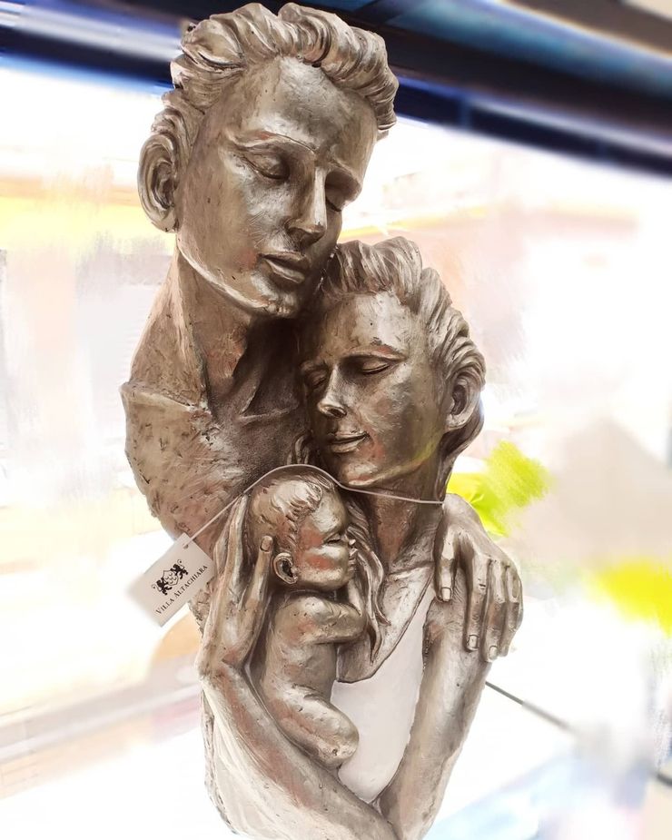 Scultura di una famiglia: un uomo con un braccio intorno alla donna che tiene in braccio un bambino, tutti e tre con gli occhi chiusi.