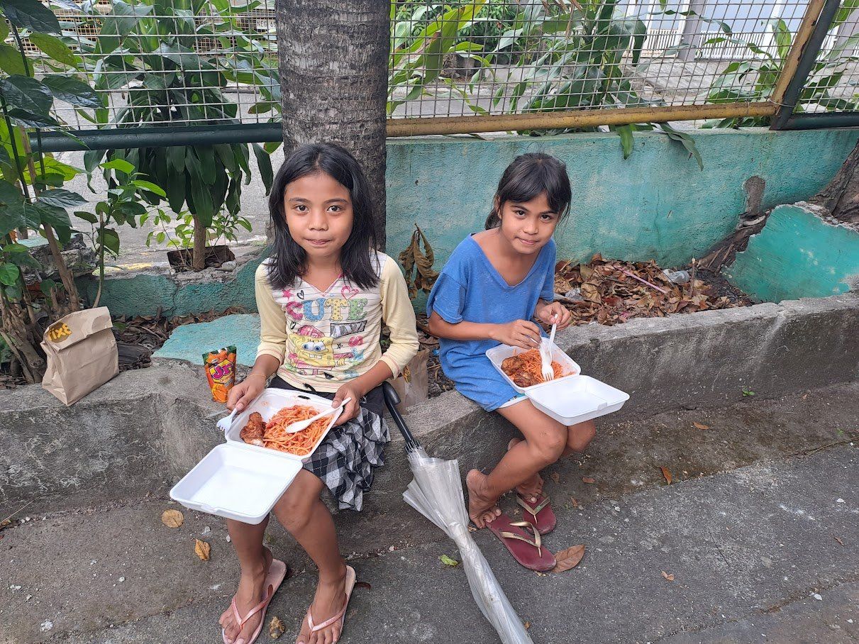 Two young girls are sitting on the sidewalk eating food.