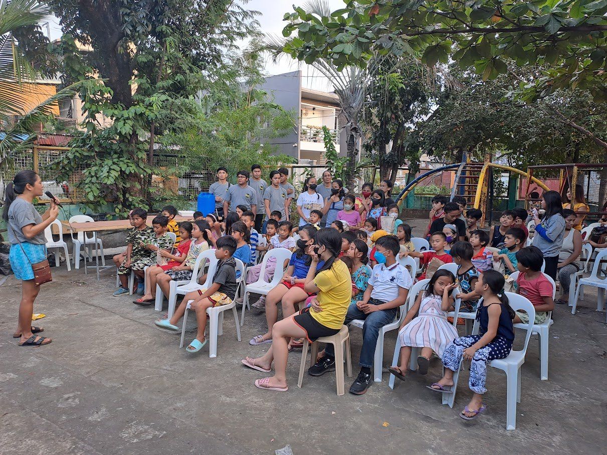 A woman is standing in front of a group of children sitting in chairs.