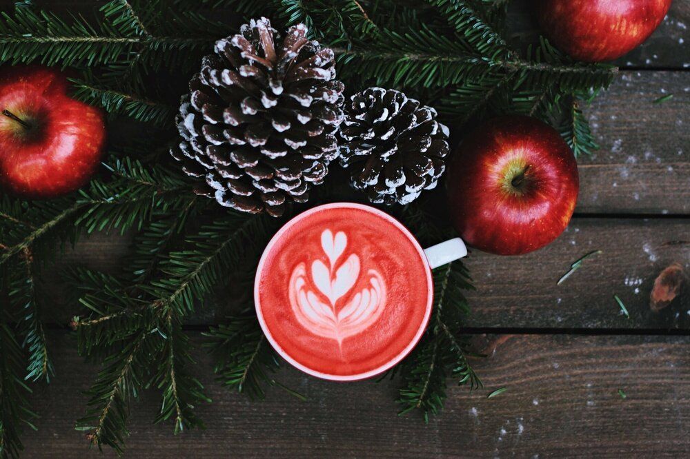 A cup of red coffee with pine cones and apples on a wooden table.