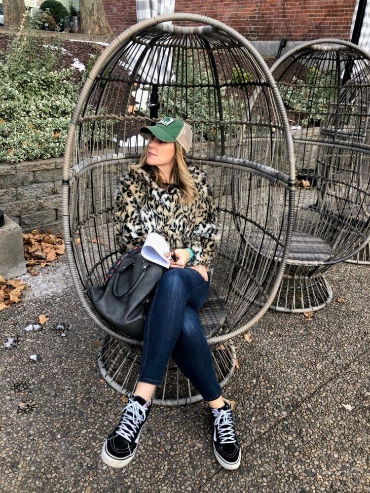 Woman in egg chair wearing hat and sneakers. Outdoors, brick path, leaves.