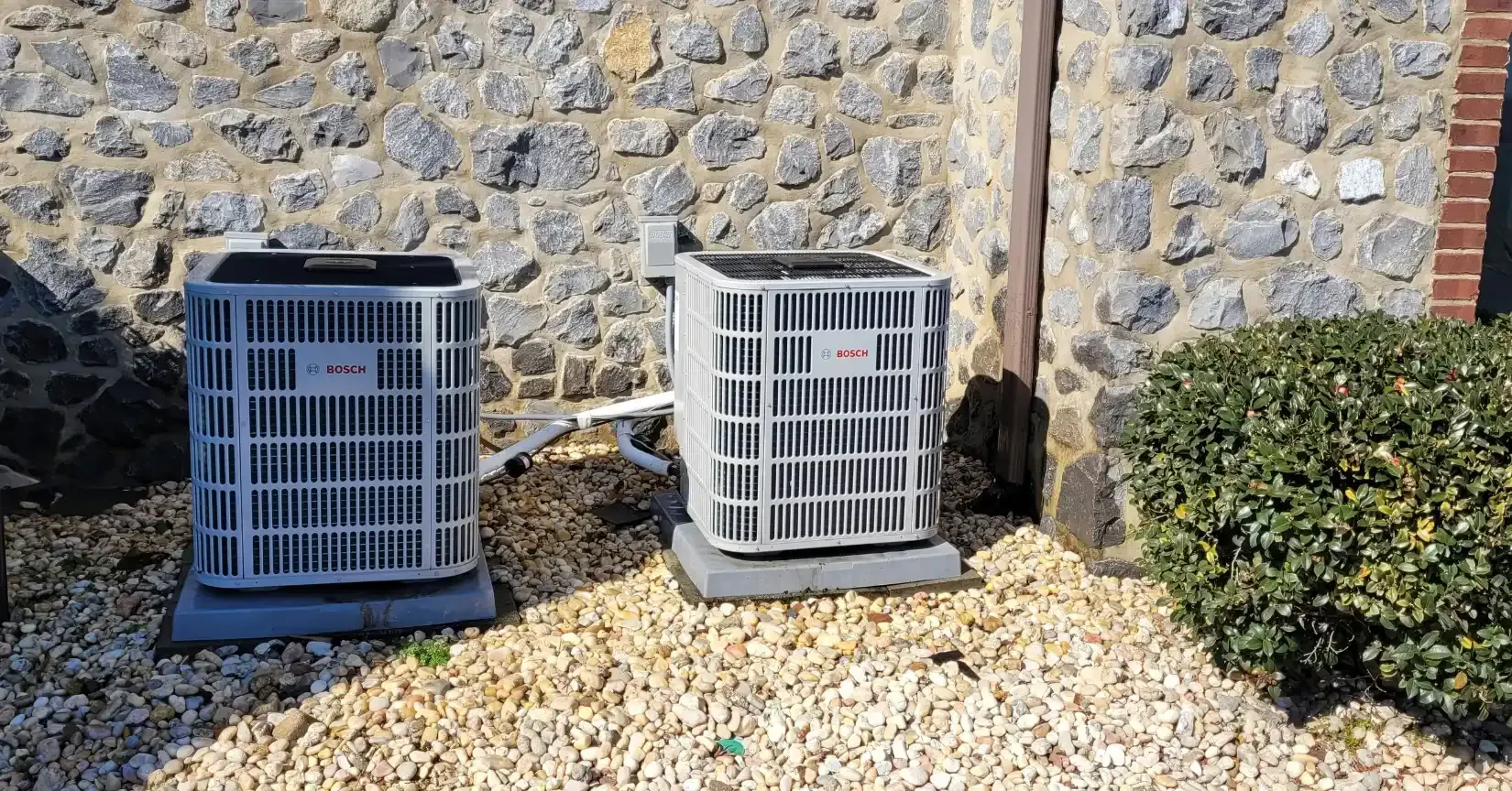 Two gray air conditioning units sitting on concrete pads, next to a stone wall.