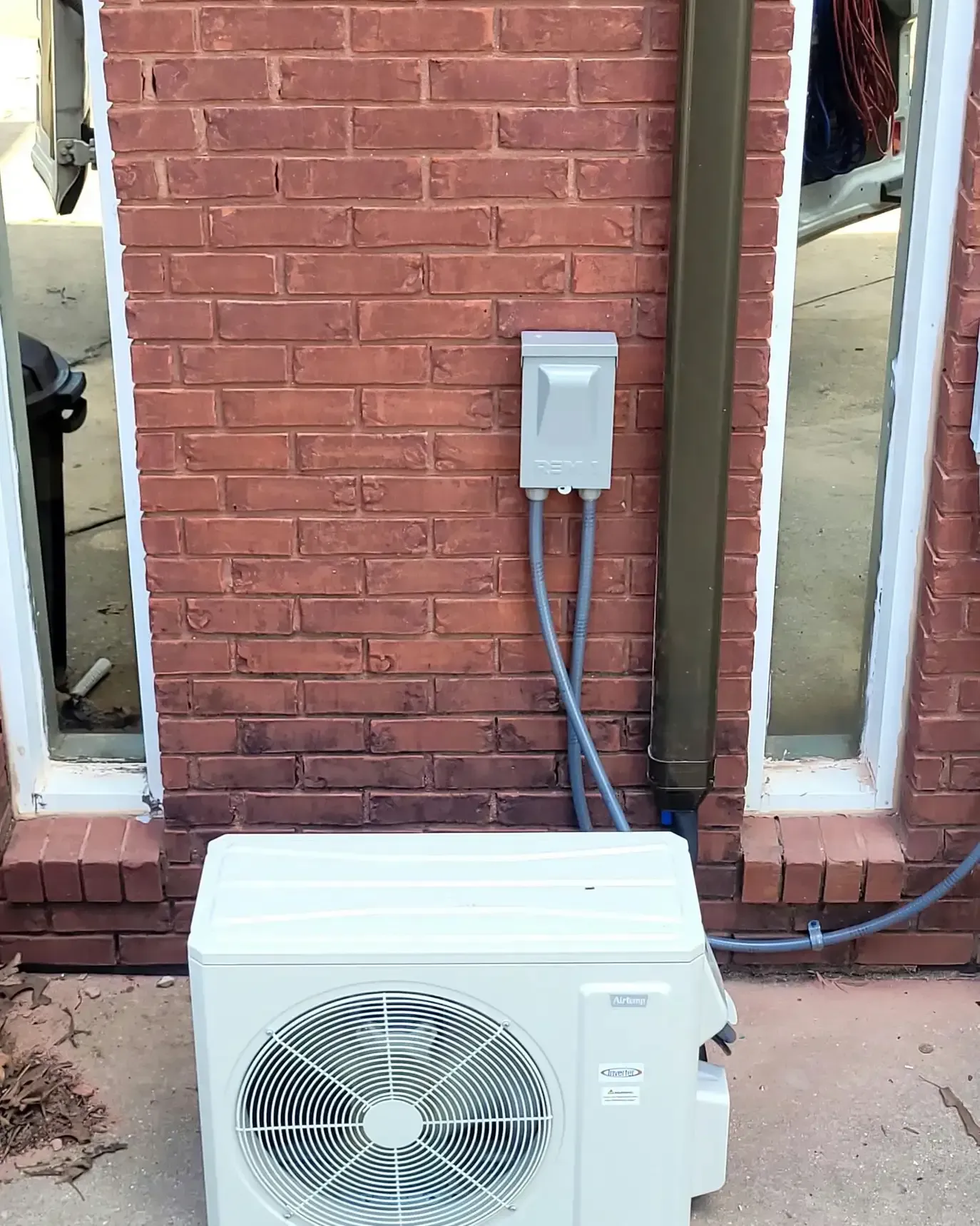 Air conditioning unit against a brick wall, below an electrical box and between two windows.