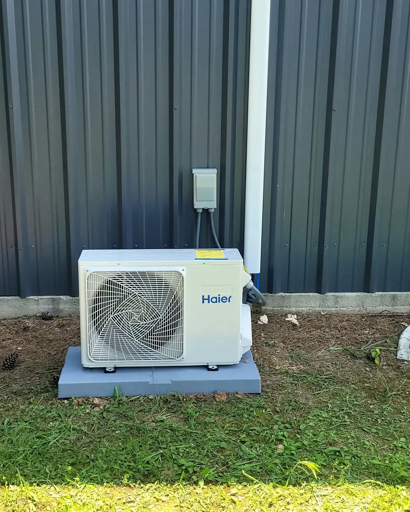 A Haier air conditioner unit on a concrete base, mounted on a grey wall with grass below.
