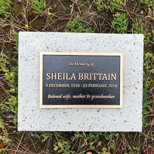 A gravestone for sheila brittain is surrounded by grass and plants.