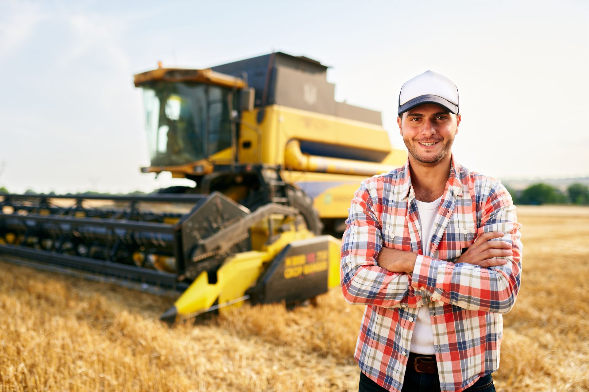 A man is standing in a field with his arms crossed in front of a combine harvester.