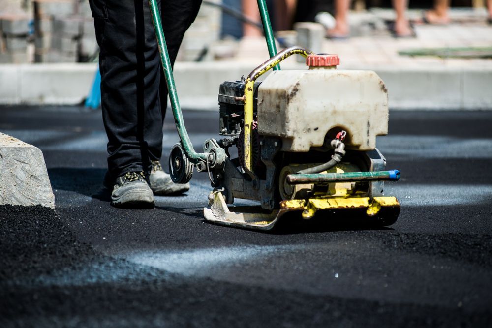 Worker Operating a Road Compactor on Fresh Black Asphalt Near Curb Stones — Lifters Hire in Cambridge, TAS