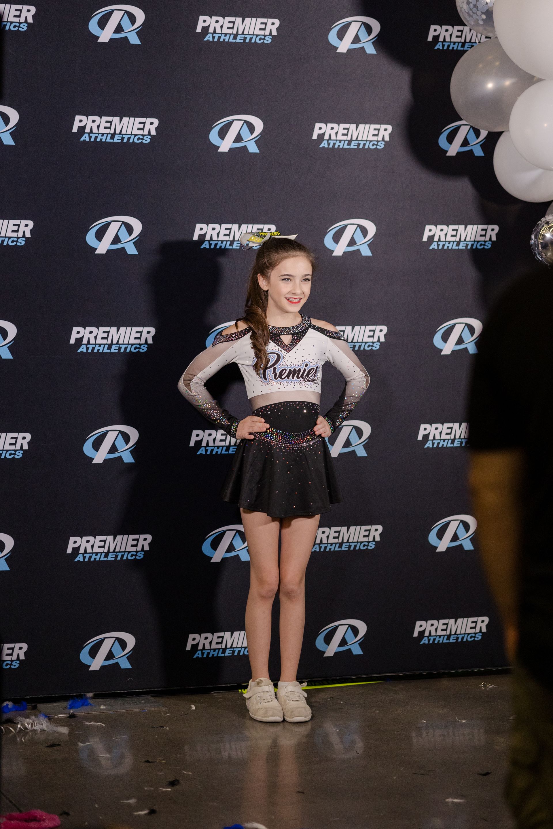 A young girl in a cheerleading outfit is standing in front of a wall with balloons.