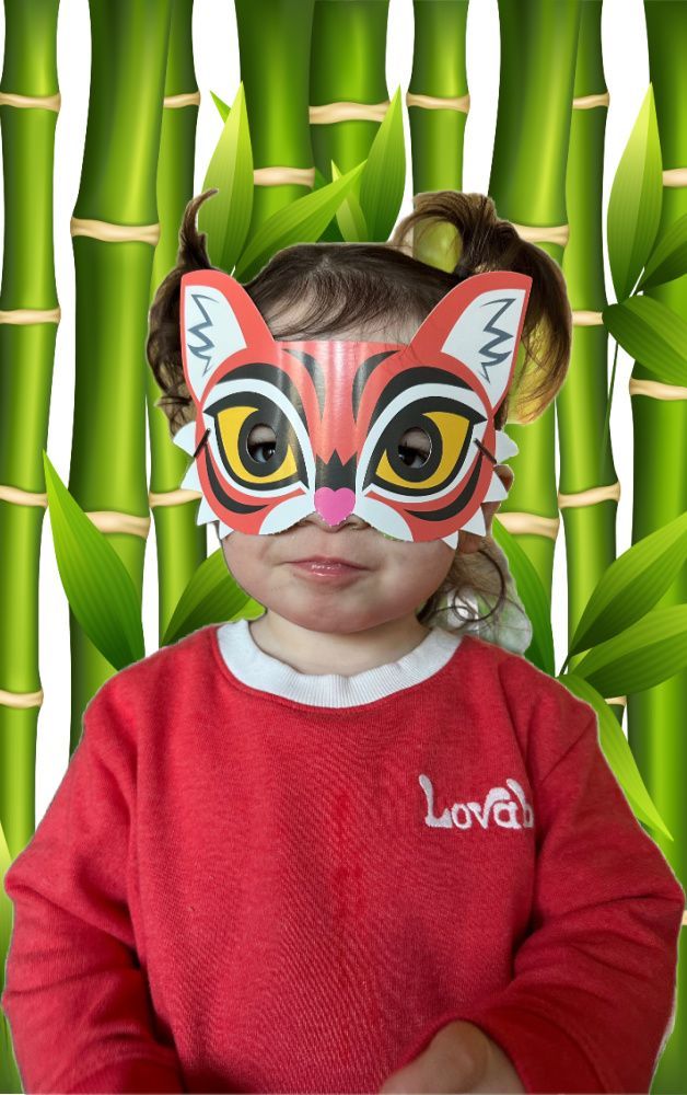 A little girl wearing a tiger mask is standing in front of a bamboo fence.