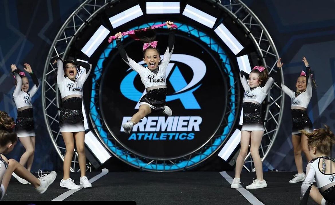 A group of cheerleaders are performing in front of a premier athletics logo