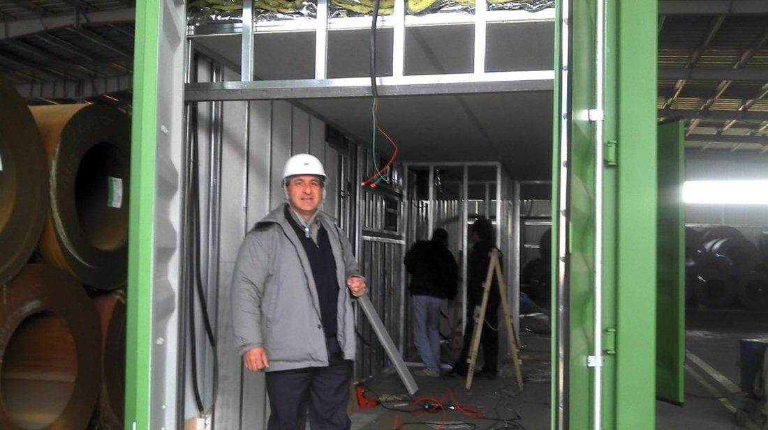 Man in hard hat stands in doorway of a construction site. Other workers and metal frames visible.