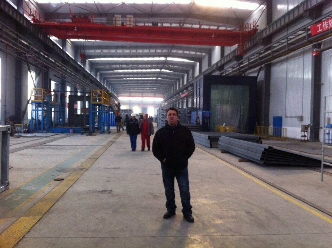 Man in black jacket stands in a large industrial building, others walking in the background.