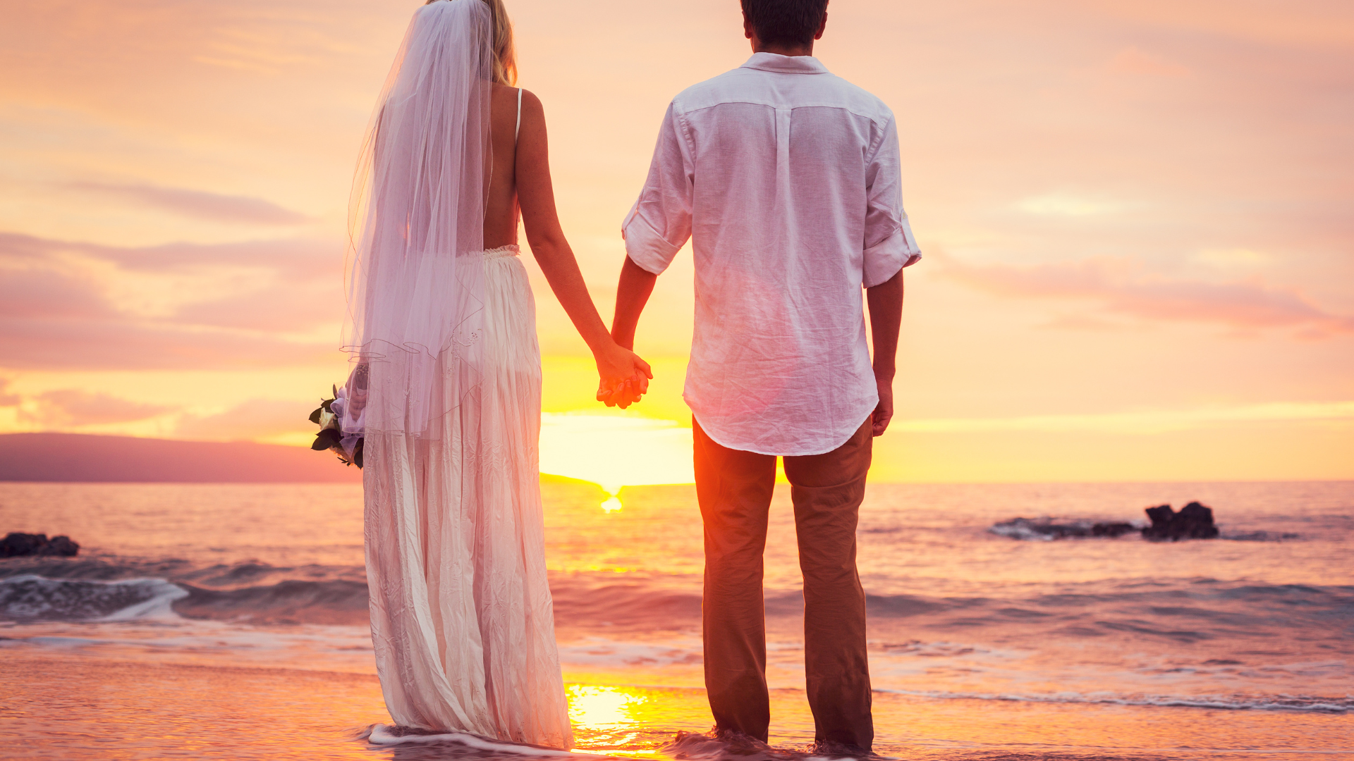 A bride and groom are holding hands on the beach at sunset.