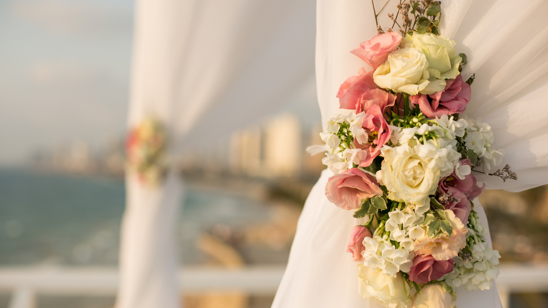 Wedding arch with white fabric, flowers and blurred ocean background.