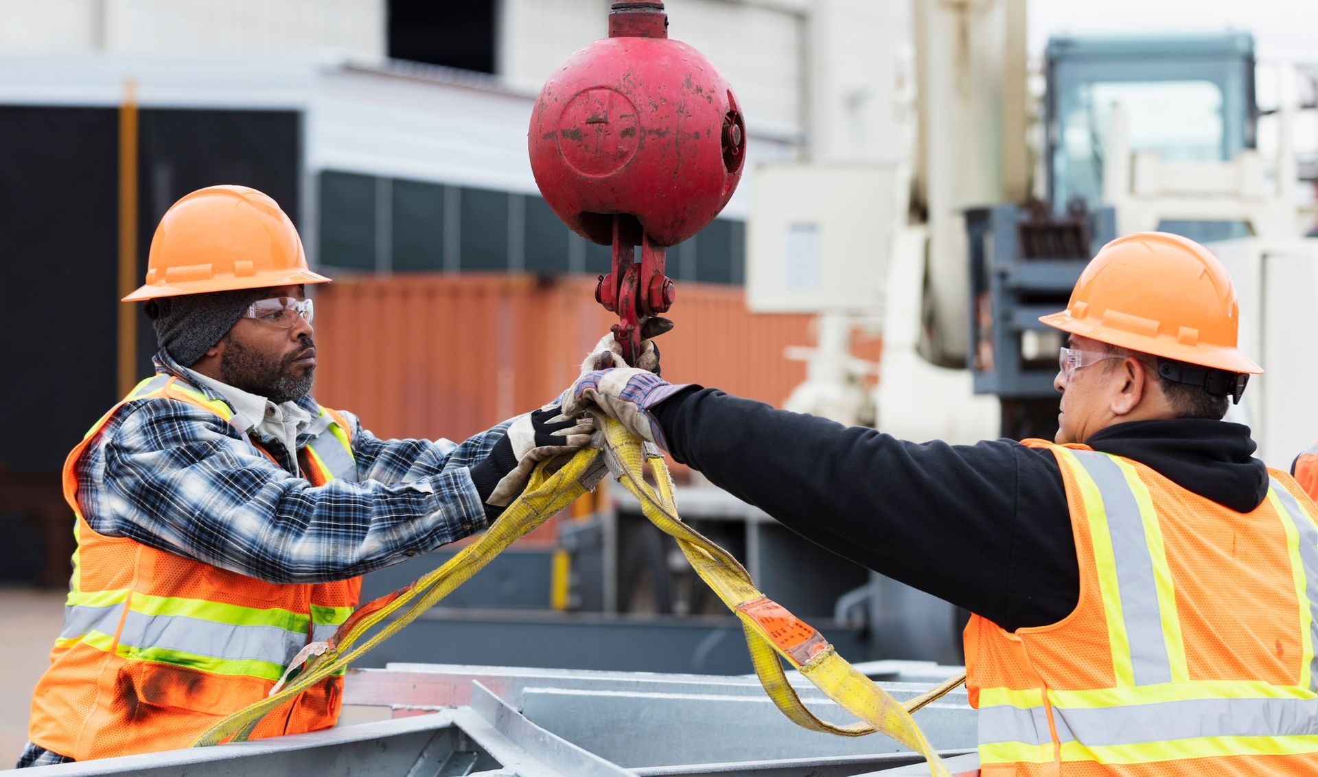 Two workers in safety gear attaching straps to a red crane hook. Two workers in safety gear attaching straps to a red crane hook.
