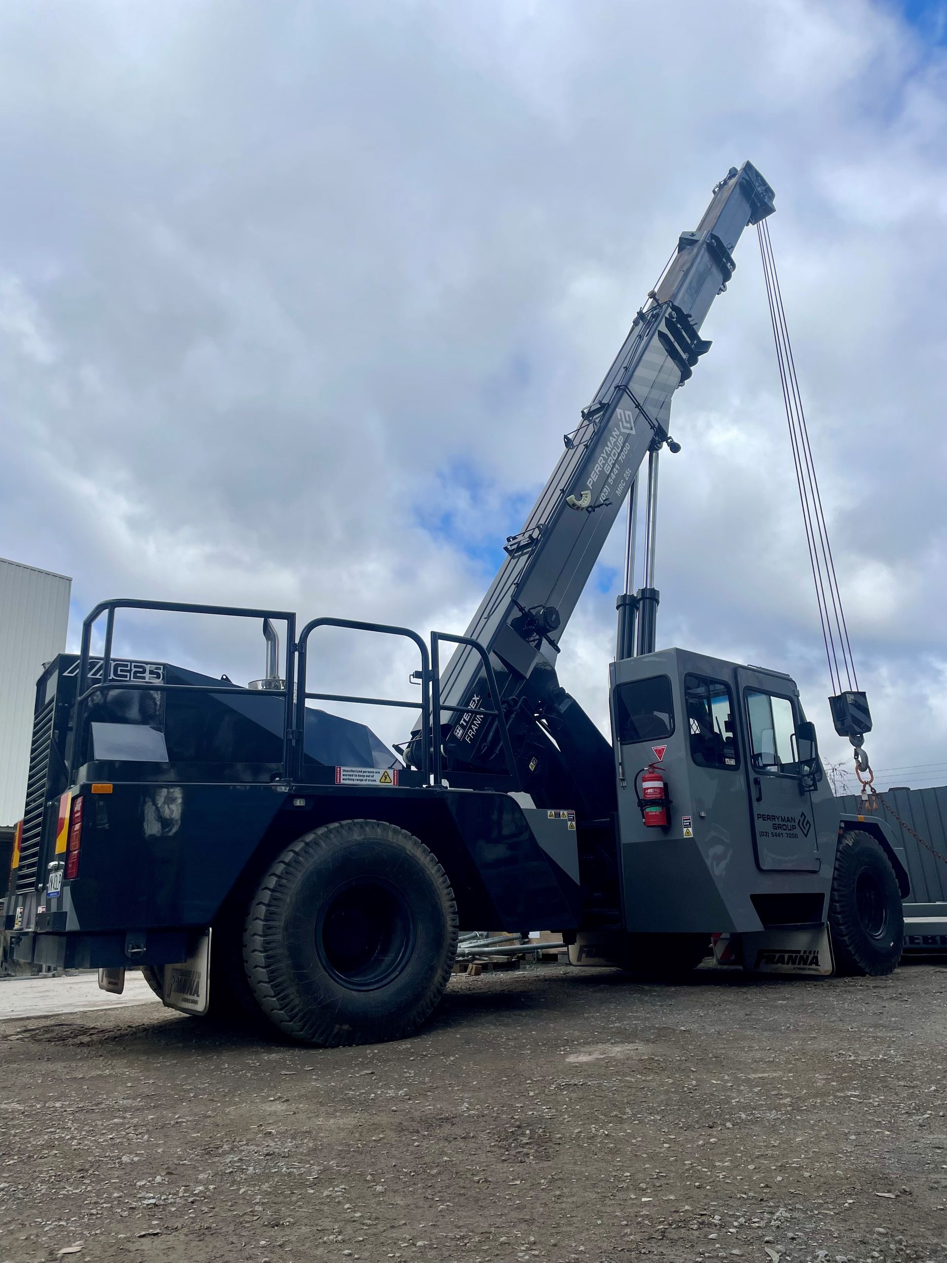 Dark grey mobile crane on a gravel lot under a cloudy sky. Its boom is extended.