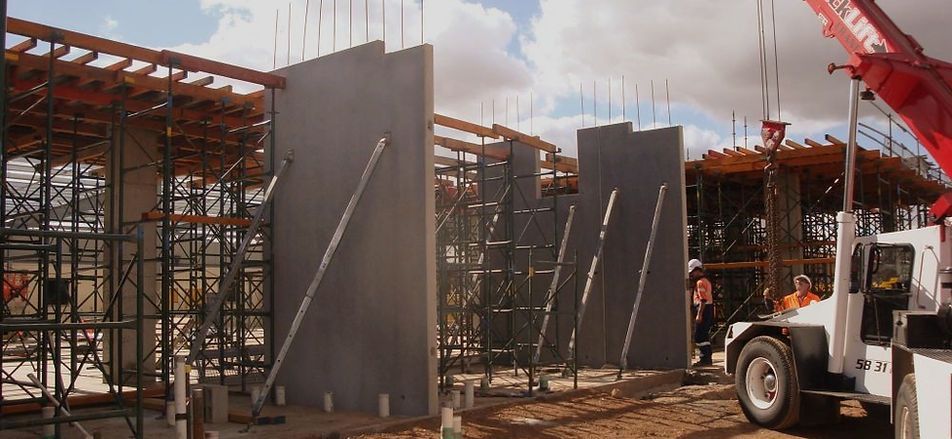 Construction site with gray concrete walls being erected by a crane; workers in the background.