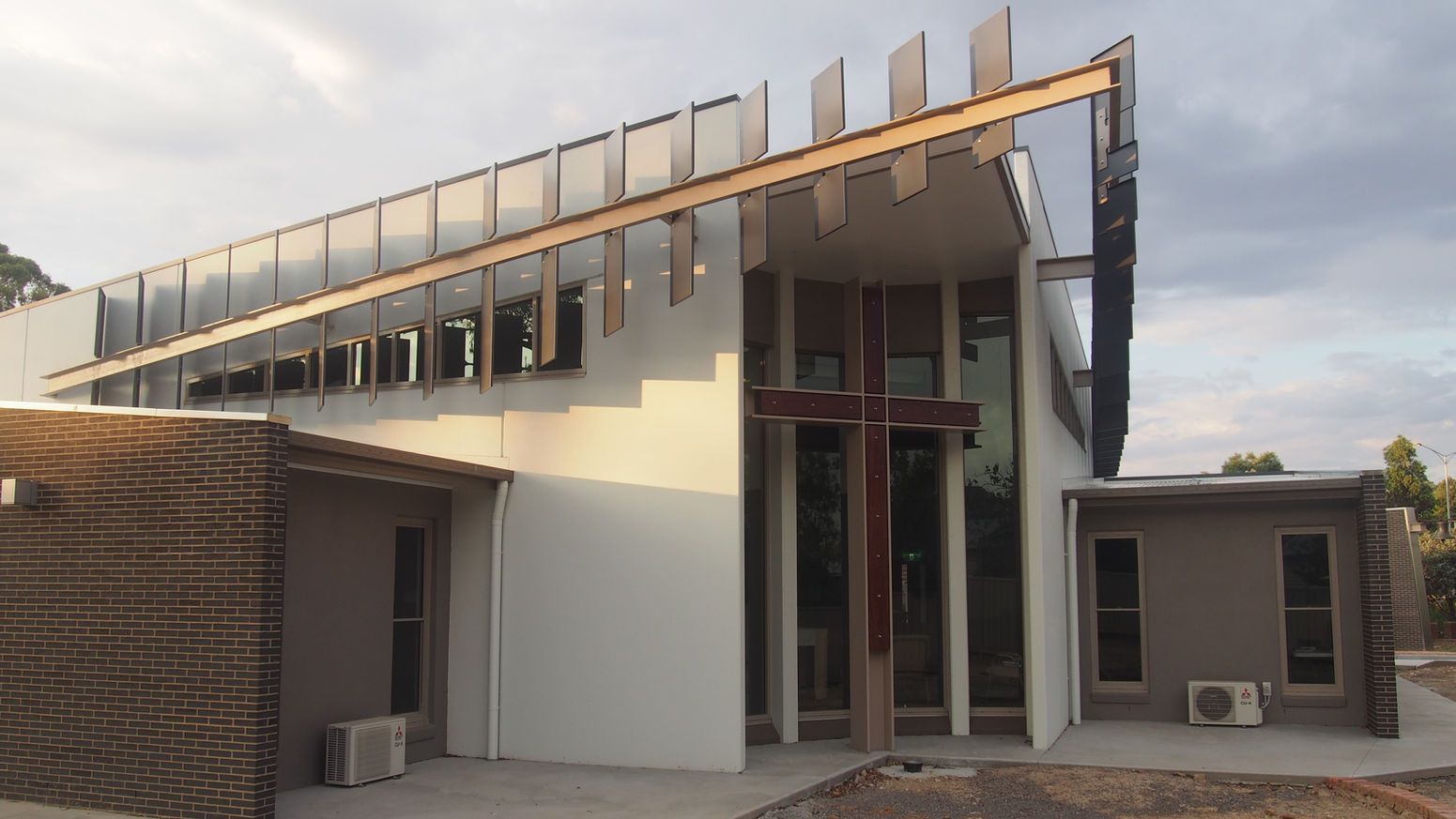 Modern church building with a large wooden cross on the facade, glass windows, and a sunlit exterior.
