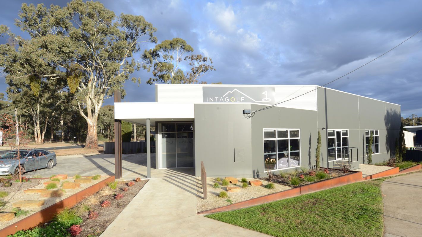 Modern gray building with white trim, glass entrance, windows, and manicured landscaping. Cloudy sky.