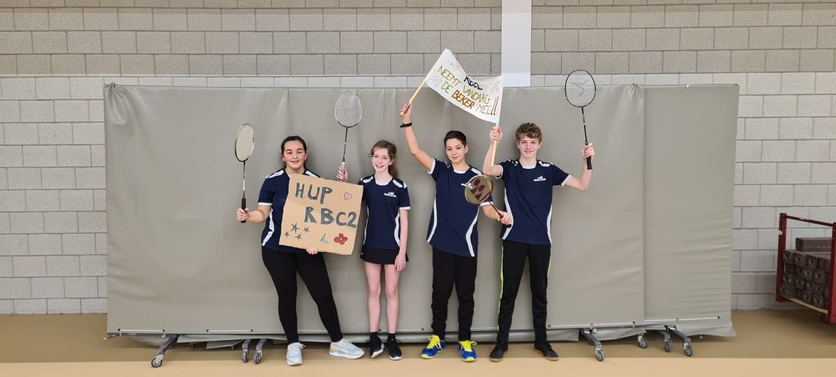 Vier jongeren in bijpassende blauwe sportkleding staan in een gymzaal met badmintonrackets, een bord en een vlag.