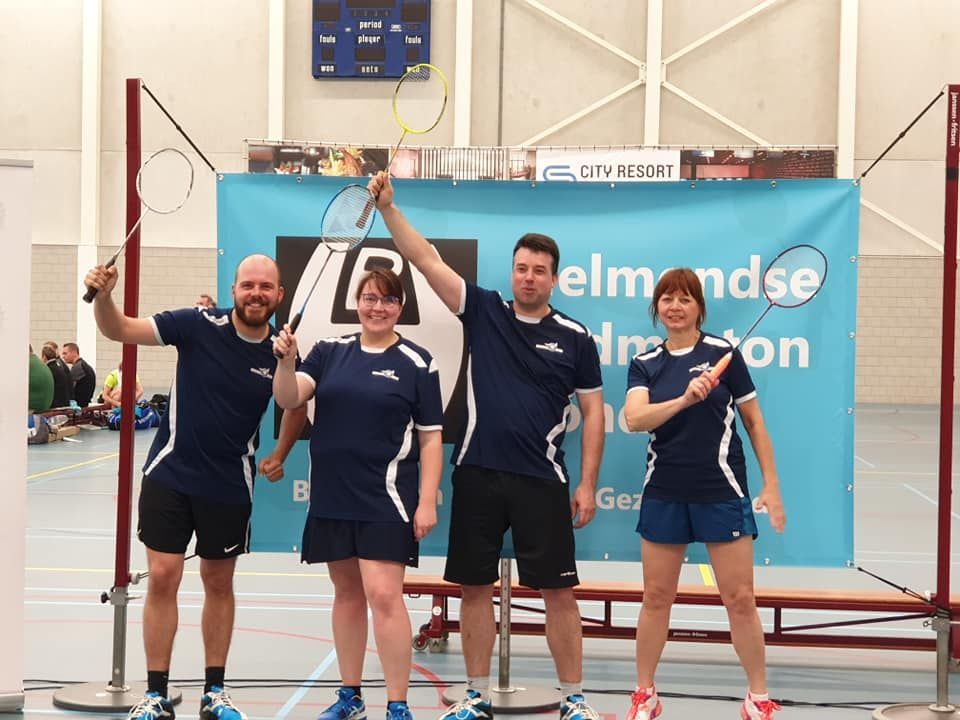 Vier personen in bijpassende blauwe badmintonuniformen poseren met rackets voor een spandoek met het logo van de sporthal.