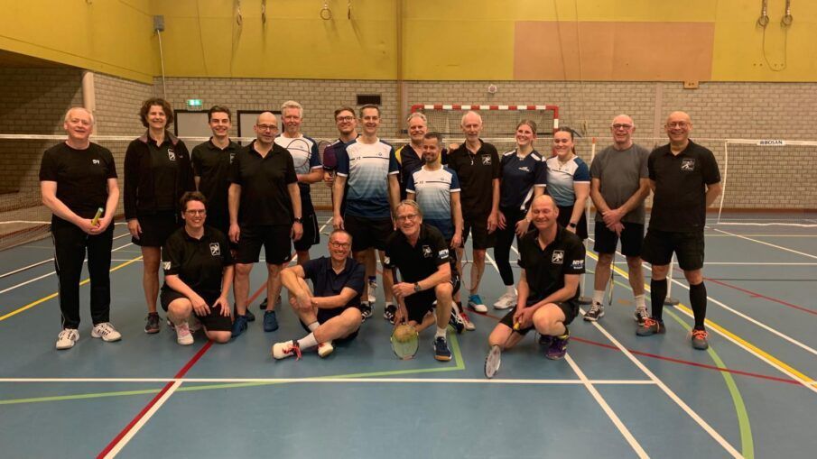 Een groep badmintonspelers poseert voor een foto op een indoorbaan, staand en knielend in twee rijen.