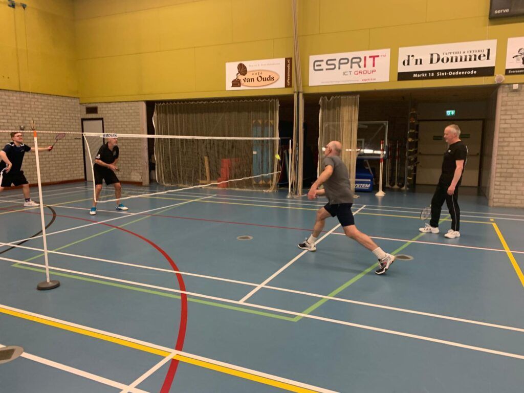 Vier mensen spelen badminton op een indoorbaan met een blauwe vloer en gele muren.