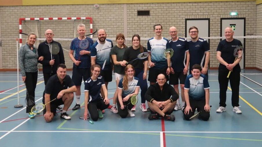 Een groep badmintonspelers poseert voor een foto op een indoorbaan, met rackets in de hand en een lachend gezicht.