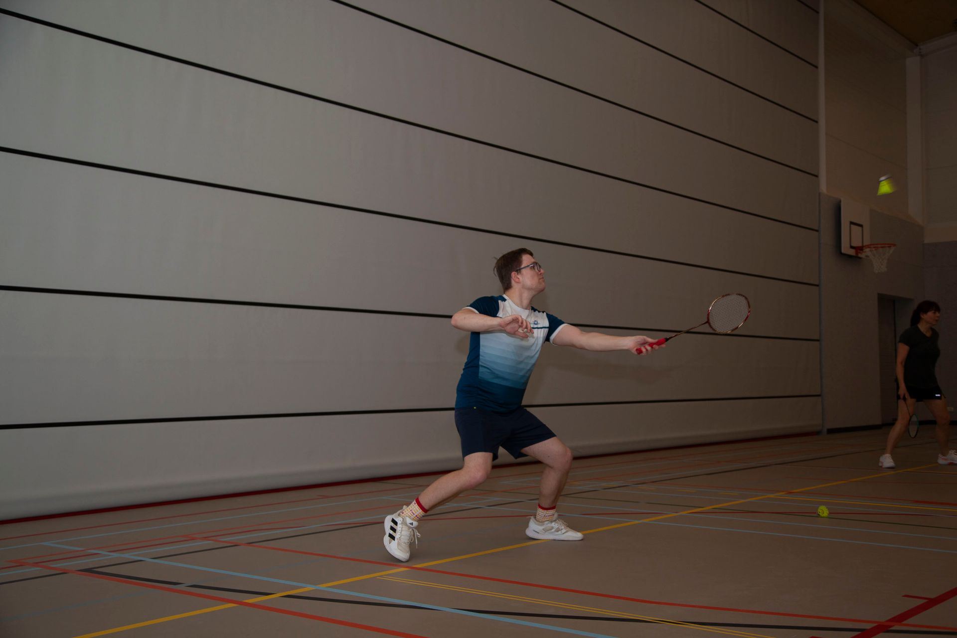 Twee mensen spelen badminton op een indoorbaan, waarbij één persoon op de voorgrond een shuttle probeert te raken.