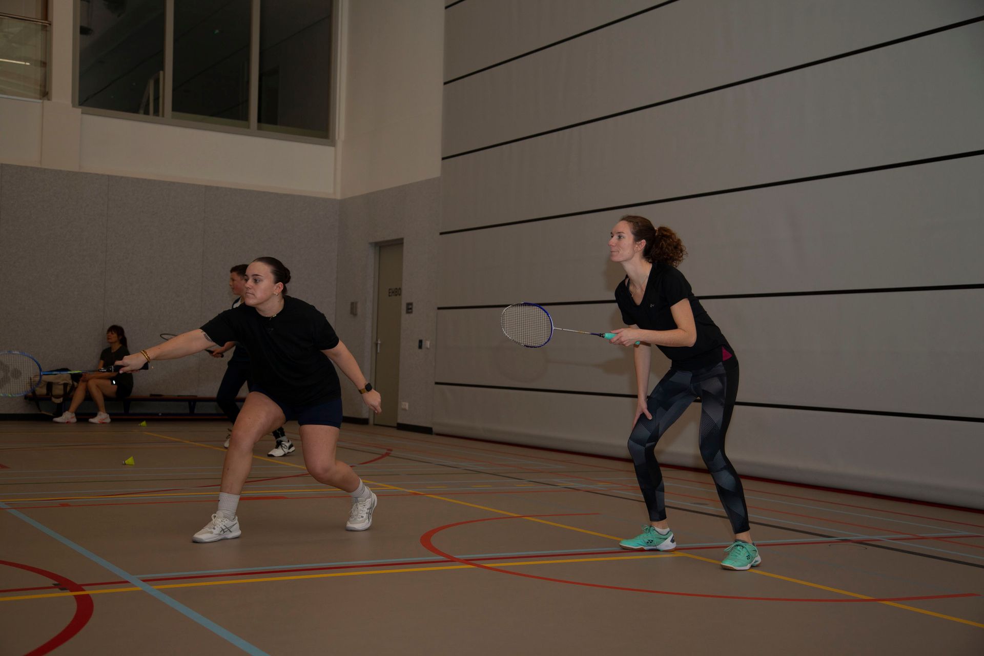 Twee mensen spelen badminton op een indoorbaan; de ene reikt naar voren om de shuttle te slaan