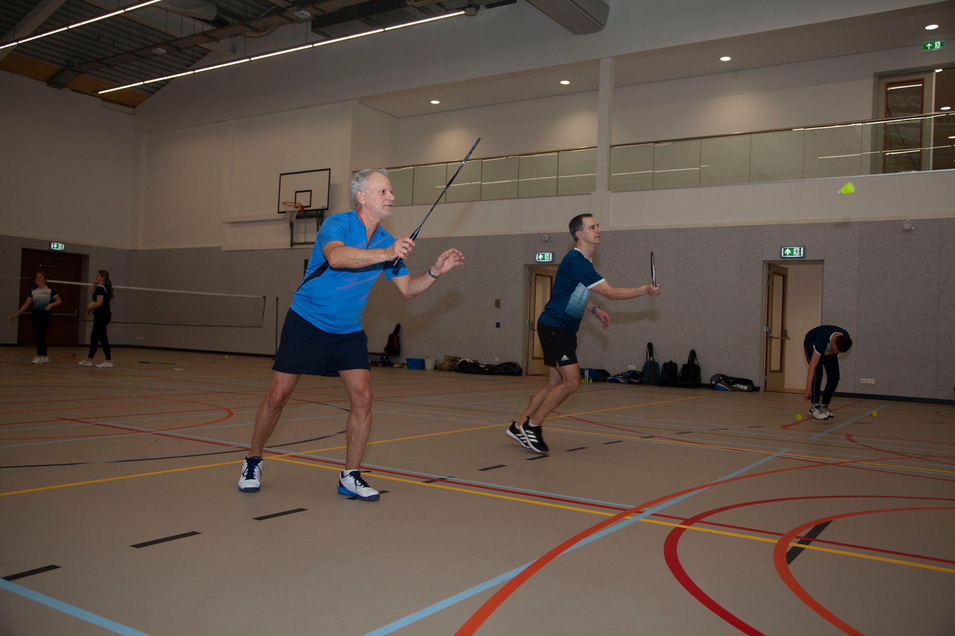 Twee mensen spelen badminton in een overdekte sporthal; de ene maakt zich klaar om de shuttle te slaan